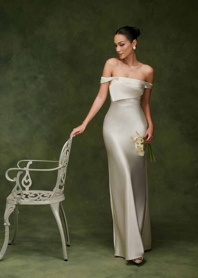 Nada off-shoulder satin dress in ivory – three-quarter view, hand resting on a white chair